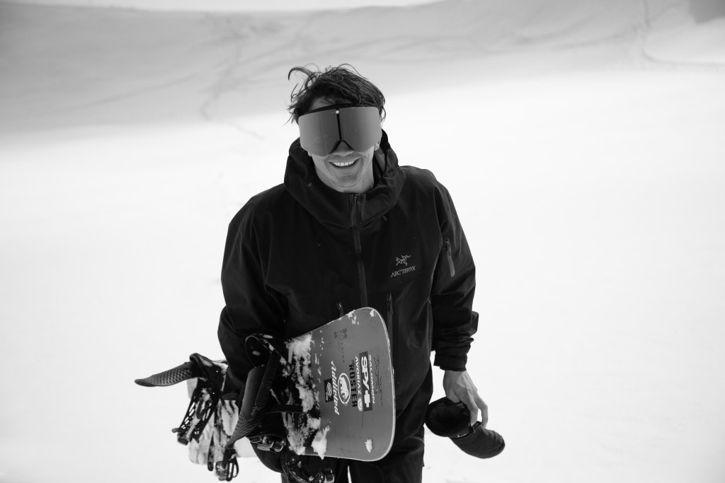 en souriant : il tient son snowboard à la main. Photo en noir et blanc.